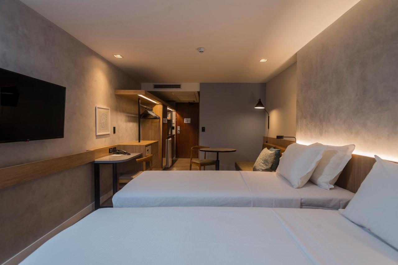 Hotel Luzeiros Fortaleza Exterior photo The photo shows a hotel room with a modern and minimalist design. There are two single beds with white linens and pillows placed next to each other. A wall-mounted flat-screen TV is positioned above a small table or desk area, which has a wooden fini