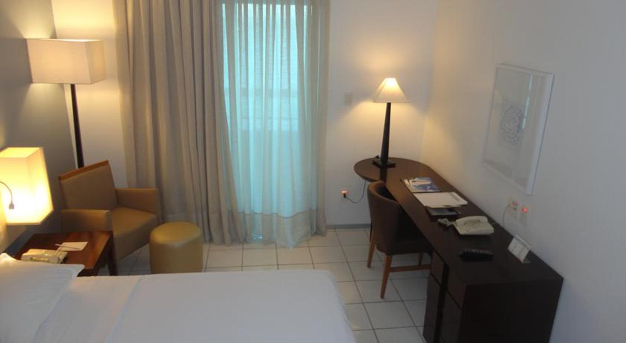 Hotel Luzeiros Fortaleza Exterior photo The photo shows a hotel room with a modern and minimalist design. It includes a bed with white linens, a small seating area with two chairs and a round ottoman, and a desk with some stationery items and a telephone. A lamp stands on the desk, providi