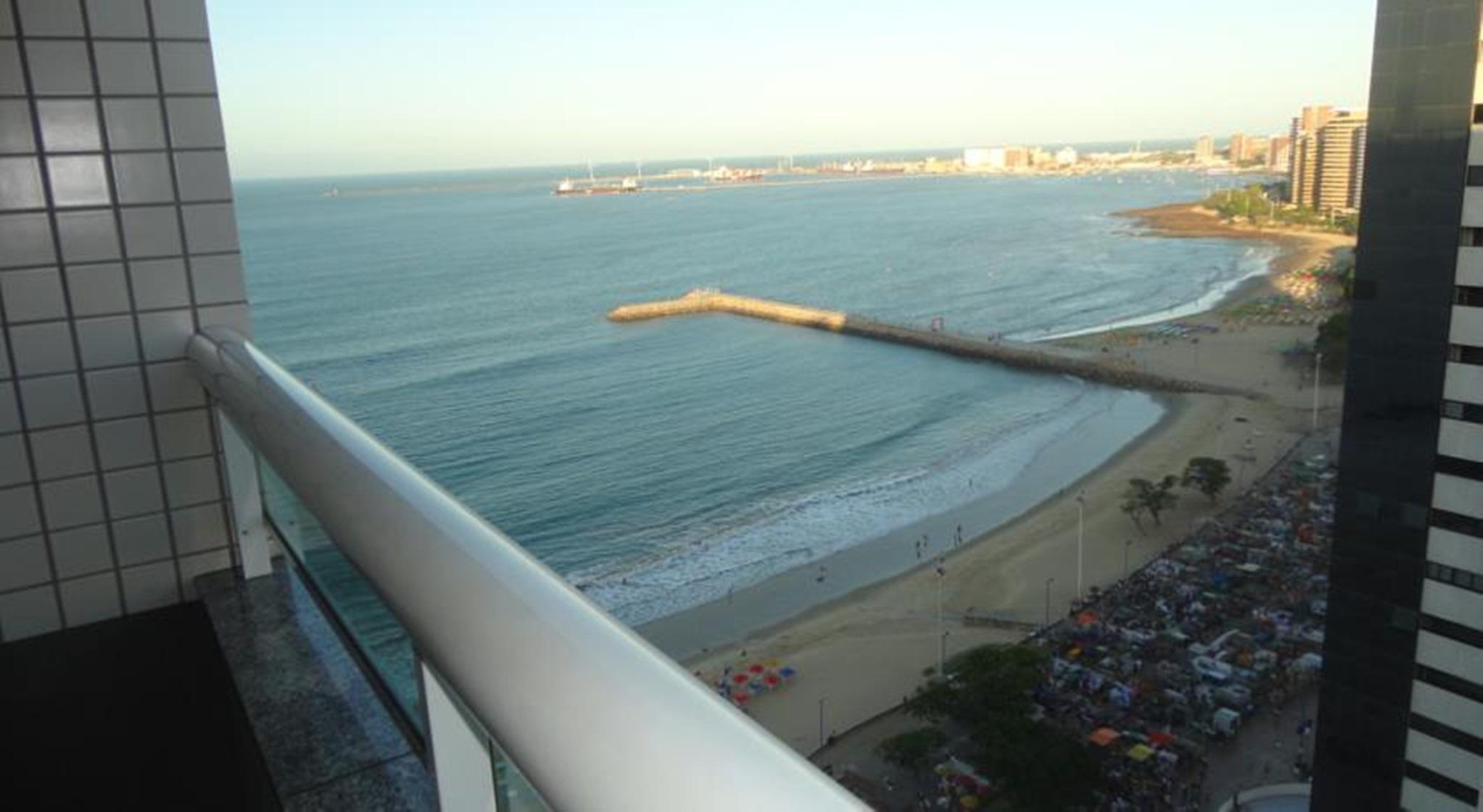 Hotel Luzeiros Fortaleza Room photo The photo depicts a coastal scene taken from a high point, possibly a balcony. In the foreground, there is a white railing, and you can see the shoreline with gentle waves lapping at the sand. There appears to be a small pier extending into the water