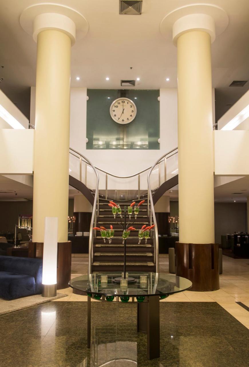 Hotel Luzeiros Fortaleza Exterior photo The photo shows an elegant interior of a hotel lobby. There is a grand, curved staircase in the center, leading to an upper level. The staircase is flanked by large columns, and there are decorative plants or floral arrangements at its base. Above th